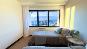 Un dormitorio con un ventanal grande, una cama, una mesita de noche con lámpara y un radiador.