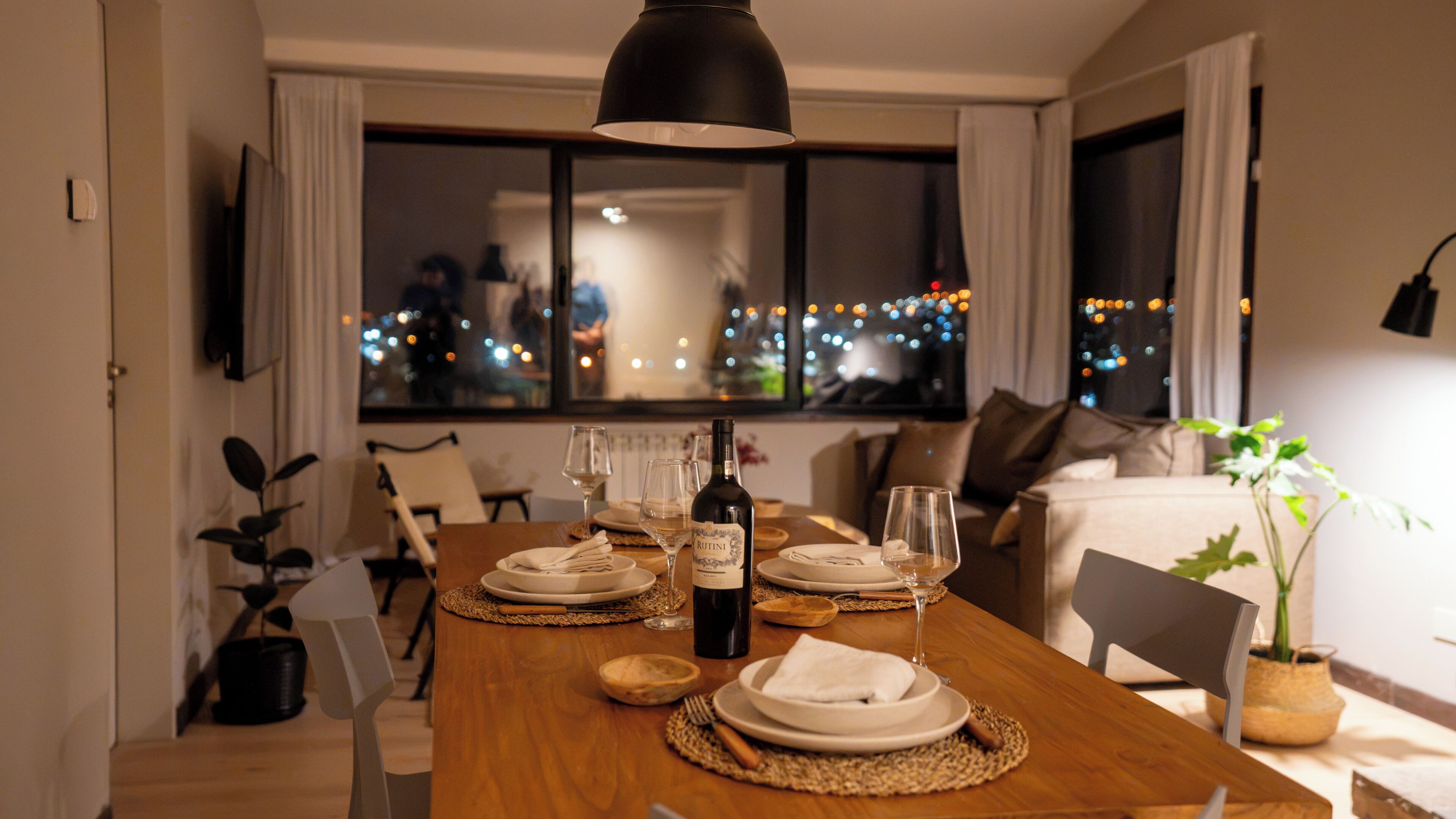 Une table à manger dressée avec des assiettes, des verres et une bouteille de vin, avec une vue sur la ville par la fenêtre.