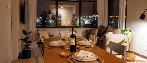 A dining table set with plates, glasses, and a bottle of wine, with a cityscape view through the window.