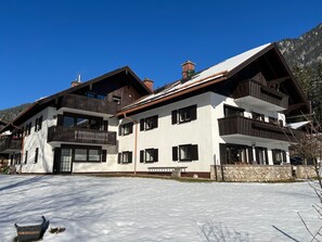 Um chalé de dois andares com varanda, uma parede de pedra e terreno coberto de neve.