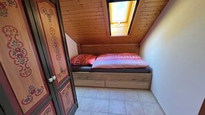 Quarto pequeno com teto inclinado, uma cama de solteiro com roupa de cama estampada e um guarda-roupa de madeira com painéis decorativos.