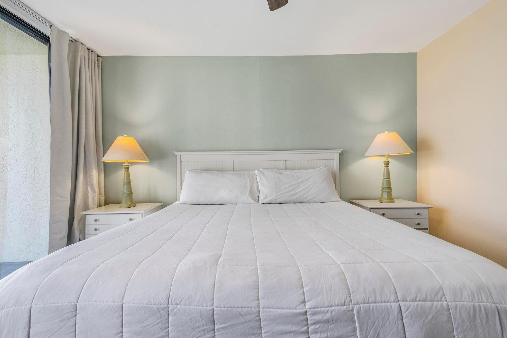 A neatly made bed with a white duvet, two bedside lamps, and a window with curtains.