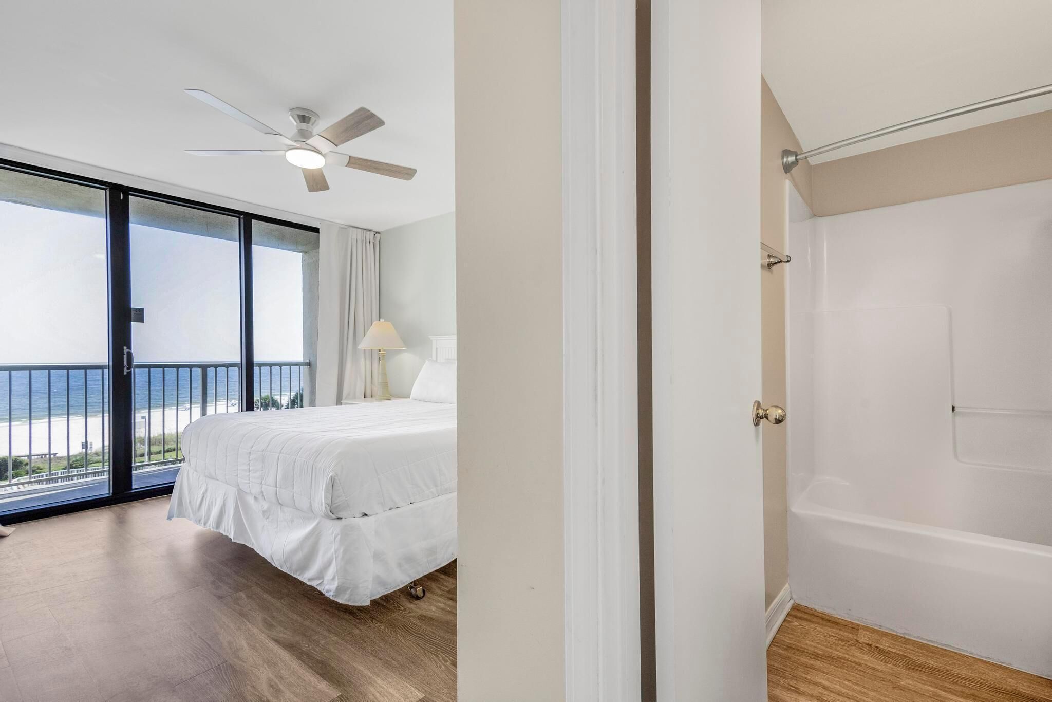 A hotel room with a bed, a ceiling fan, a view of the sea, and a door leading to a bathroom.