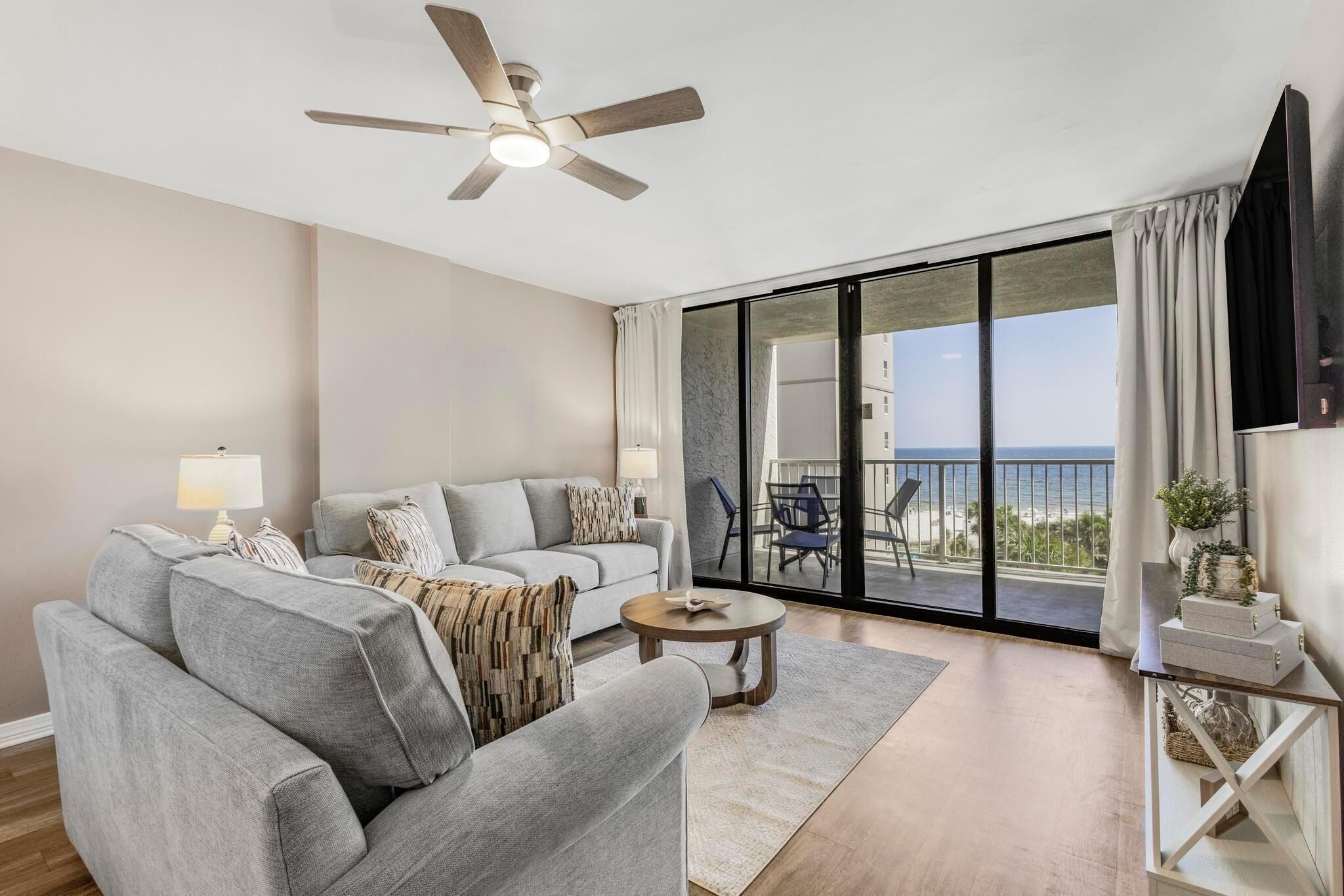 Een moderne woonkamer met een grijze bank, een salontafel en een plafondventilator. Er is een balkon met uitzicht op de oceaan.