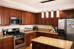Cozinha moderna com armários de madeira, eletrodomésticos de aço inoxidável, bancadas de granito e iluminação pendente.