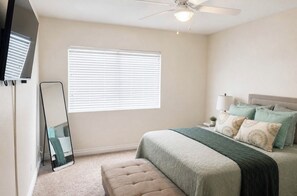 Ein Schlafzimmer mit Bett, Deckenventilator, Fenster mit Jalousien, Spiegel und Nachttisch mit Lampe.