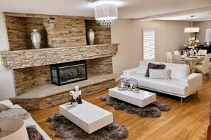 A living room with a stone fireplace, white sofas, and a dining area with a chandelier. - Modern 4BR Pool & Spa Oasis • Sleeps 10 (Las Vegas)