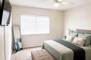 Un dormitorio con cama, espejo de pie, ventilador de techo y ventana con persianas.