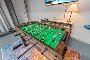 A foosball table in a room with a television mounted on the wall, a lamp, and a door. - Game Room, Pool & Fire Pit Near Madeira Beach (Seminole)