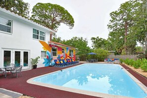 A swimming pool with lounge chairs and umbrellas, surrounded by a house with a colorful mural. - Game Room, Pool & Fire Pit Near Madeira Beach (Seminole)
