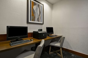 A hotel room with a desk, two monitors, a printer, a keyboard, and two chairs.