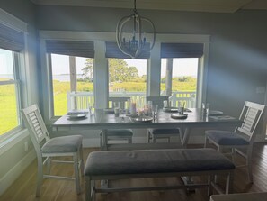 A dining area with a view of a grassy field and a body of water, featuring a table set with plates, glasses, and a candle. - MarshMellow -Peaceful Knott's Island Waterfront Retreat (knotts island)