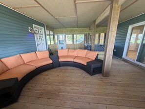 A covered outdoor seating area with a curved sectional sofa, wooden deck flooring, and a view of the surrounding landscape.