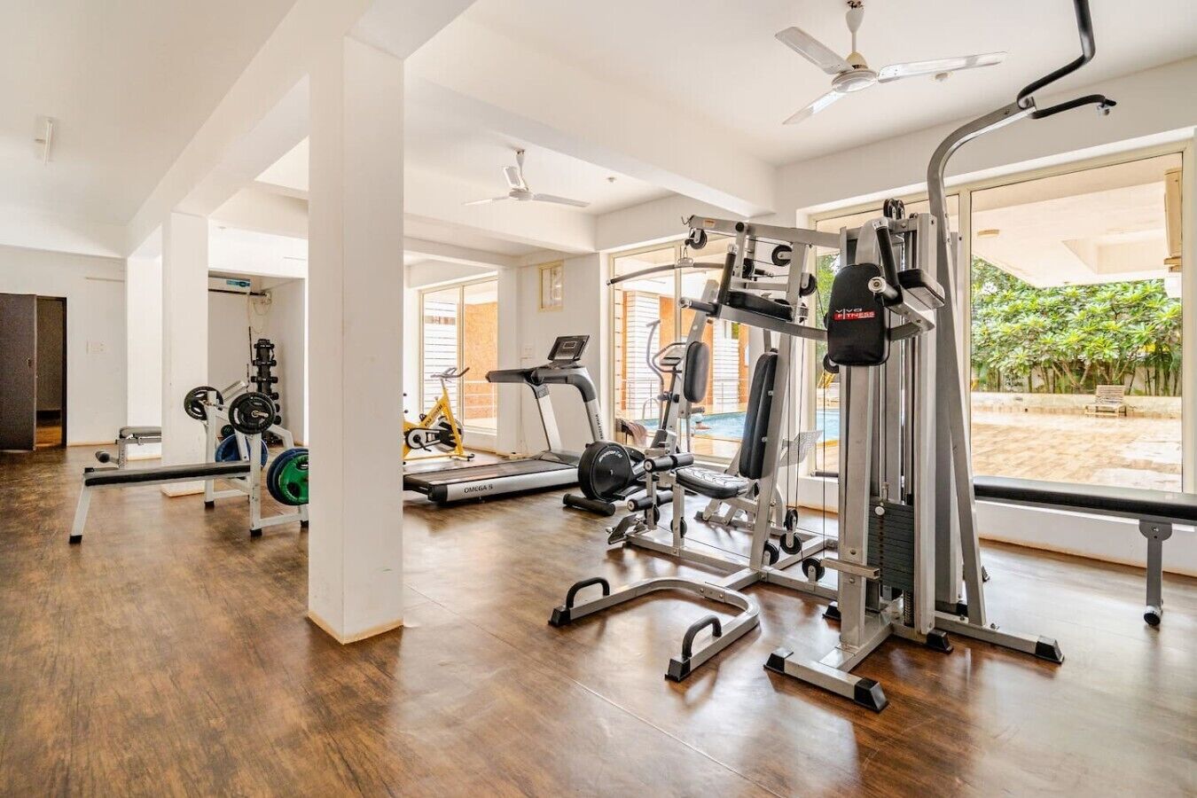 Ein Fitnessraum mit unterschiedlichen Trainingsgeräten, einem Laufband und Blick auf einen Innenhof.