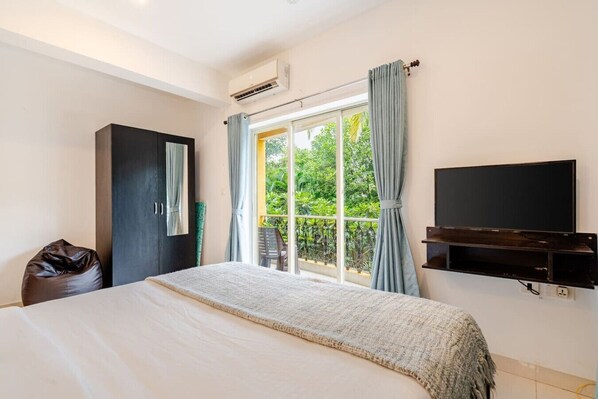 A hotel room with a bed, a TV mounted on the wall, a window with curtains, and an air conditioning unit.