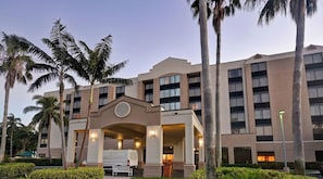 Ein mehrstöckiges Hotel mit zentralem Eingang, Palmen und einem gepflegten Rasen.