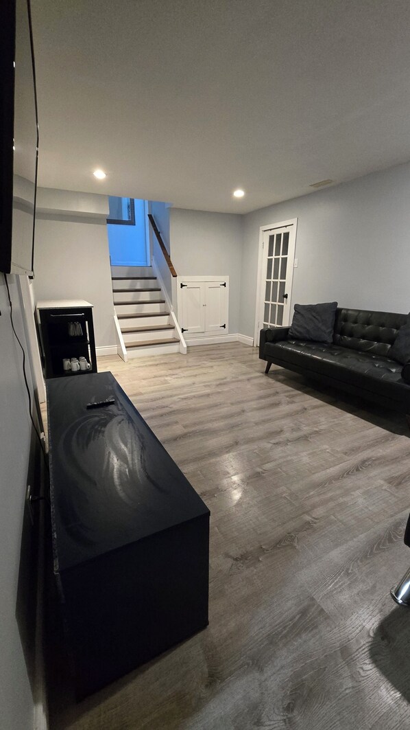 A spacious living area with a staircase leading to an upper floor, a black ottoman, a leather sofa, and a television mounted on the wall.