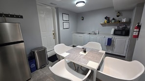 A compact kitchen with a stainless steel refrigerator, a sink, and a microwave. There is a dining table with two chairs and a small trash bin.