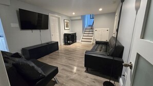 A modern living room with a flat-screen TV, a black sofa, a black leather couch, and a staircase leading to a second floor. - Modern comfort meets unique two level split suite. Family size and Pet Friendly (Cobourg)