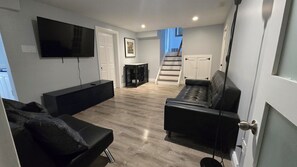 A modern living room with a flat-screen TV, a black sofa, a black leather couch, and a staircase leading to a second floor. - Modern comfort meets unique two level split suite. Family size and Pet Friendly (Cobourg)