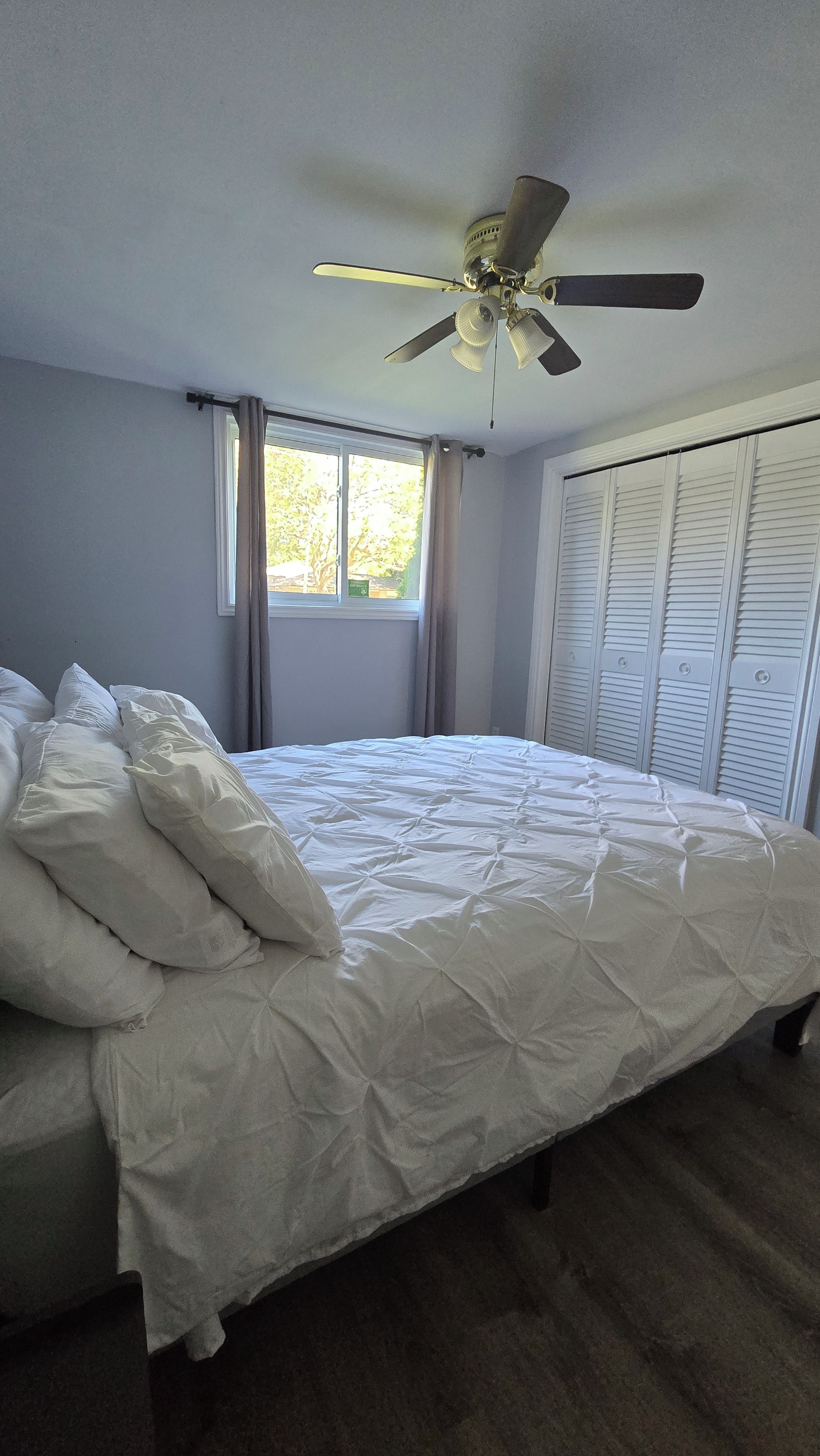 Ein Schlafzimmer mit einem Bett, einem Deckenventilator, einem Fenster mit Vorhängen und einer Reihe von Klappschranktüren.