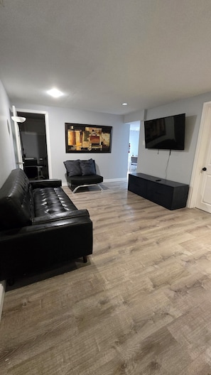 A modern living room with a black leather sofa, a flat-screen TV mounted on the wall, and a wooden floor.