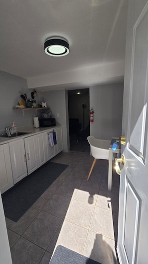 A kitchen with white cabinets, a microwave, and a sink. There is a white chair and a door with a key in the lock.