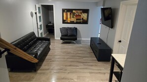 A modern living room with a black leather sofa, a black armchair, a wooden staircase, a flat-screen TV, and a painting on the wall. - Modern comfort meets unique two level split suite. Family size and Pet Friendly (Cobourg)