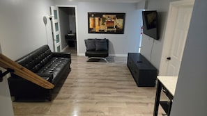 A modern living room with a black leather sofa, a black armchair, a wooden staircase, a flat-screen TV, and a painting on the wall. - Modern comfort meets unique two level split suite. Family size and Pet Friendly (Cobourg)
