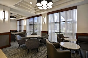 A hotel lobby with round tables, cushioned seating, large windows with sheer curtains, and modern pendant lighting.