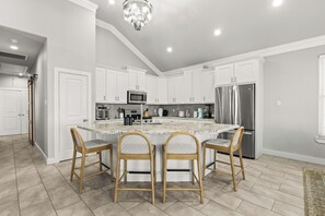 Una cocina moderna con armarios blancos, una isla central con taburetes altos, electrodomésticos de acero inoxidable y una araña.
