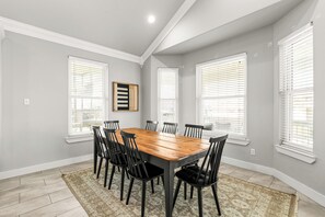 Uma sala de jantar com mesa de madeira e cadeiras pretas, grandes janelas com persianas e piso em tons claros.