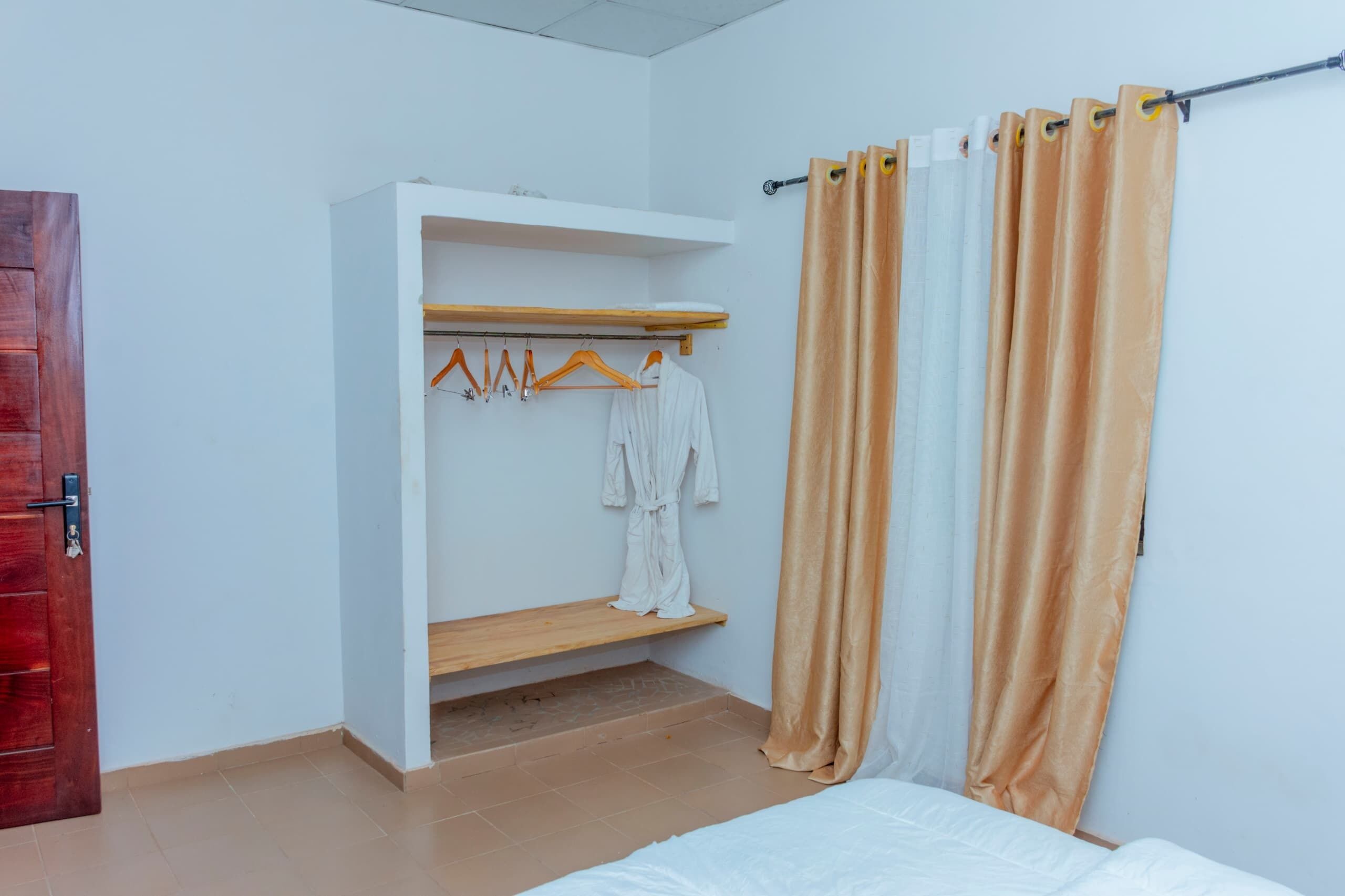 A room with a bed, a wooden shelf with hangers, and a white robe hanging on the wall.
