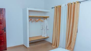 A room with a bed, a wooden shelf with hangers, and a white robe hanging on the wall.