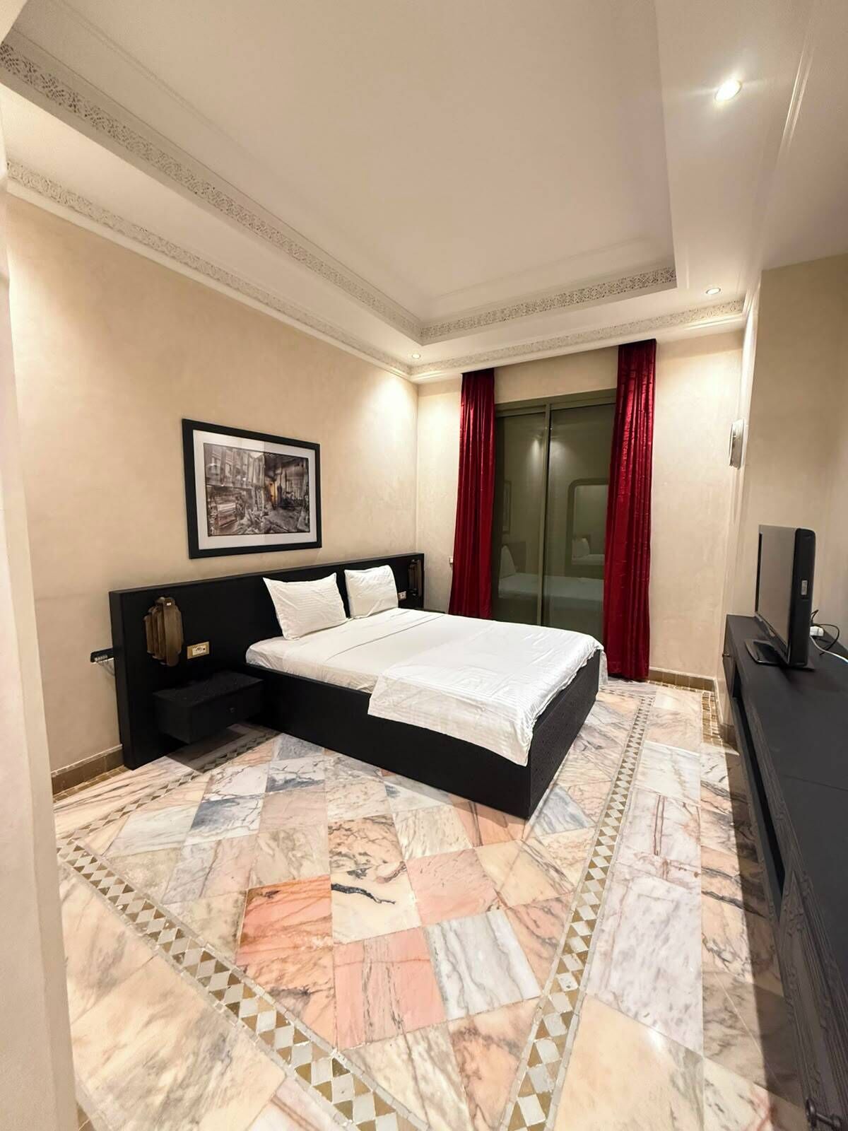 A hotel room with a bed, a television, a painting on the wall, and a patterned floor.