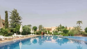 A clear swimming pool with lounge chairs and umbrellas, surrounded by greenery and a building in the background. - BELLA Palmeraie Apartment (Marrakech)