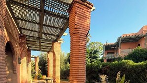 A covered outdoor area with brick columns, a tiled floor, and a garden with plants and a potted tree. - BELLA Palmeraie Apartment (Marrakech)