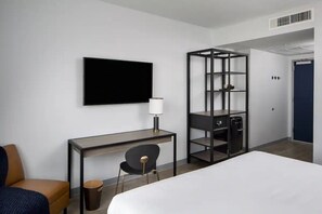 Ein Hotelzimmer mit Bett, Schreibtisch mit Stuhl, Fernseher, Lampe, Mülleimer und Regale.