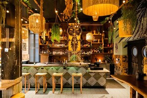 A bar with a patterned floor, wooden stools, hanging lights, and shelves stocked with bottles.