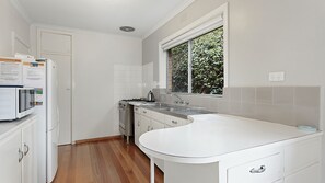 A modern kitchen with white cabinetry, a built-in oven, a sink, and a window with a view of greenery. - Bright Beach House - Point Lonsdale (POINT LONSDALE)