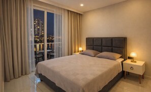 A hotel room with a large bed, two bedside tables with lamps, a view of a cityscape through floor-to-ceiling windows, and a balcony with a table and chairs. - Royal Grand Exculisive luxury apartment in charming Alanya (Alanya)