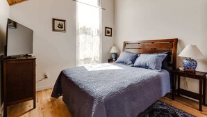 Une chambre avec un plafond en bois, un lit recouvert d’une courtepointe bleue, deux lampes de chevet, un téléviseur posé sur un meuble en bois et une fenêtre ornée de rideaux légers.