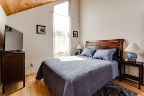 A bedroom with a wooden ceiling, a bed with a blue bedspread, two bedside lamps, a TV on a wooden stand, and a window with sheer curtains. - 2BR + Loft Space | 5 Min to Autzen & River (Springfield)