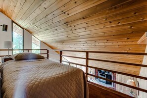 A bedroom with a wooden ceiling, a bed with a quilted cover, a nightstand, and a window with a view of a fireplace.