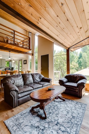A spacious living area with a high ceiling, wooden beams, a leather sofa, a wooden coffee table, and a large window offering a view of the outdoors. - 2BR + Loft Space | 5 Min to Autzen & River (Springfield)