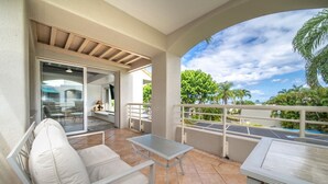 Un balcon avec un canapé blanc, une petite table et une porte vitrée donnant sur un espace intérieur comprenant une chaise et une lampe.