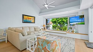 Un salon moderne comprenant un canapé, une table basse, un pouf à motifs, un ventilateur de plafond, une grande fenêtre donnant sur l’extérieur et un téléviseur.
