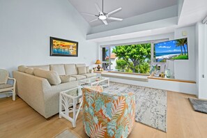 Ein modernes Wohnzimmer mit einer Couch, einem Couchtisch, einem gemusterten Ottomanen, einem Deckenventilator, einem großen Fenster mit Ausblick und einem Fernseher.