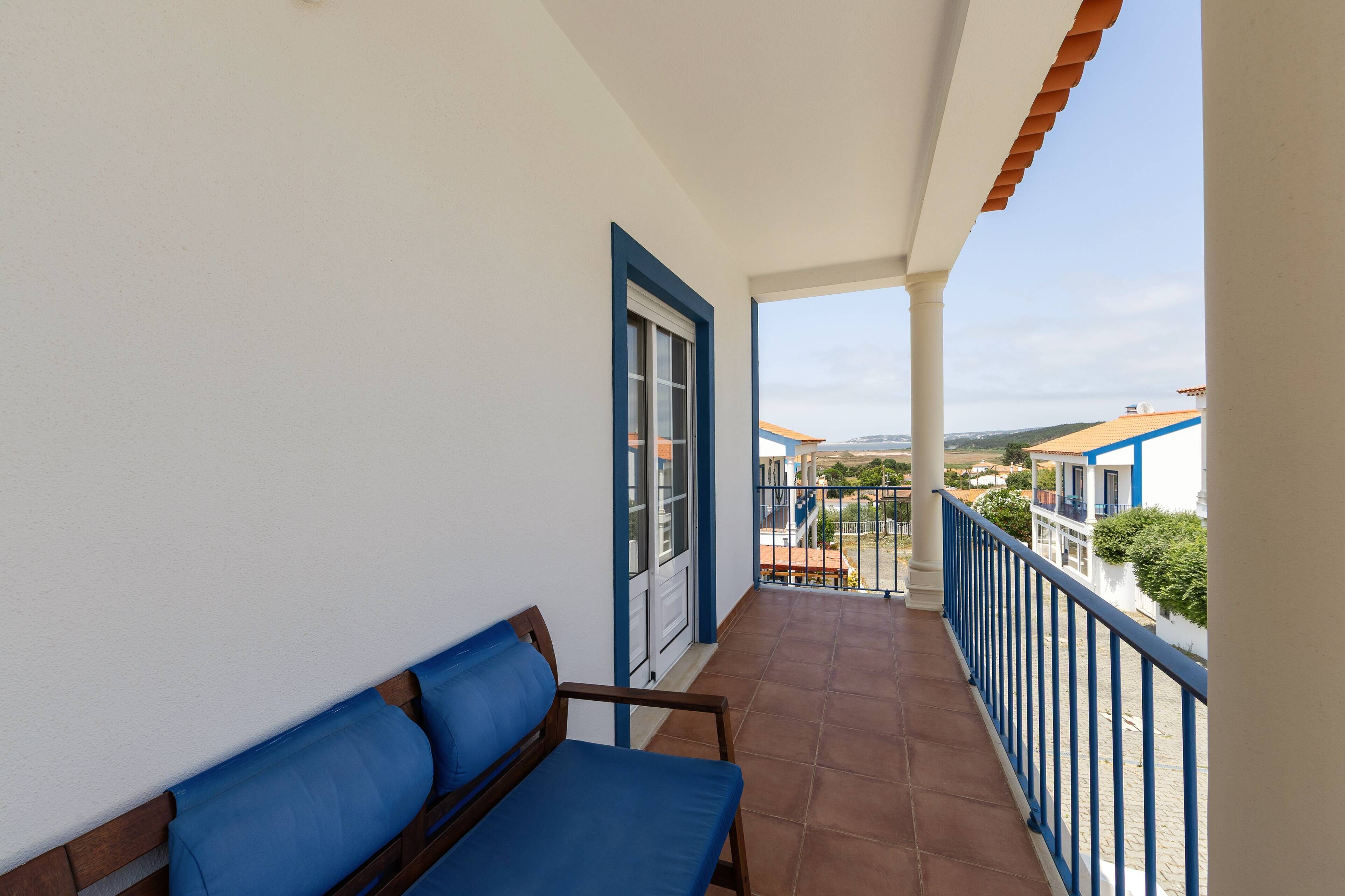 A balcony with a blue cushioned bench, a door, and a view of a coastal area with buildings.