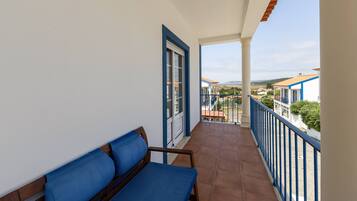 A balcony with a blue cushioned bench, a door, and a view of a coastal area with buildings.